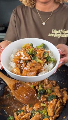 a woman is holding a bowl full of chicken and broccoli over rice with a wooden spoon