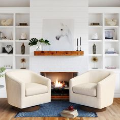 two white chairs sitting in front of a fire place with books on the shelves behind them