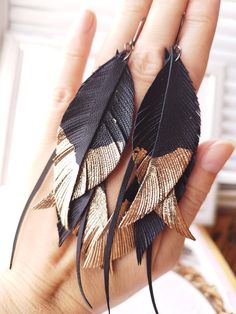 a pair of black and gold feather earrings on someone's hand with their fingers in the air