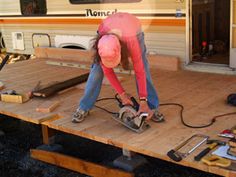 a woman in pink shirt and blue jeans working on wooden table next to camper