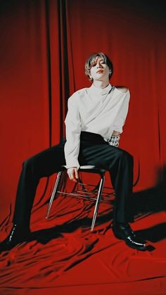 a man sitting on top of a chair in front of a red curtain with his legs crossed