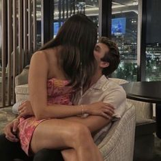 a man and woman sitting on a couch in front of a window with the city lights behind them