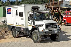 a white truck parked in front of a building with other trucks behind it on a dirt road