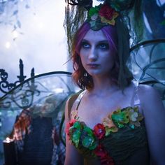 a woman with pink hair and flowers on her head standing in front of a wrought iron fence