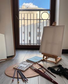 an easel, pencils, and other art supplies sit on the floor in front of a window