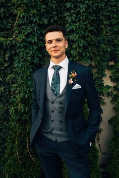 a man wearing a suit and tie standing in front of a wall with green plants