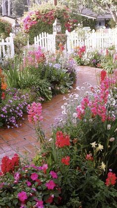 a garden filled with lots of colorful flowers next to a white picket fence and trees