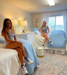 two women sitting on beds in a bedroom with blue sheets and white walls, one is smiling at the camera