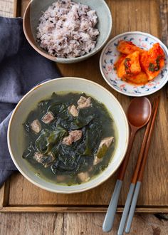 two bowls filled with meat and greens next to some chopsticks on a wooden tray