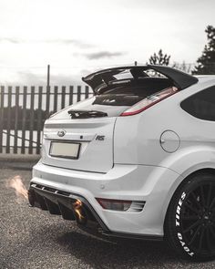 the rear end of a white car parked in a parking lot next to a fence