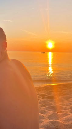 a man sitting on top of a sandy beach next to the ocean under a sunset