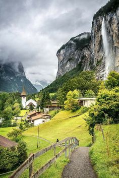 a path leading to a small village with a waterfall in the background on a cloudy day