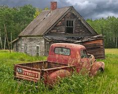 an old red truck is parked in the grass next to a run down house and barn