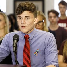 a man wearing a red tie sitting in front of a microphone with other people behind him