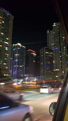 cars driving down the road in front of tall skyscrapers at night with lights on