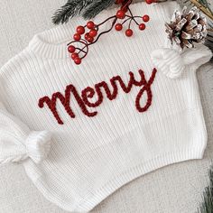 a white sweater with the word merry written on it next to pine cones and berries