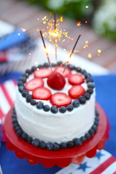 a white cake with strawberries and blueberries on top is topped with sparklers