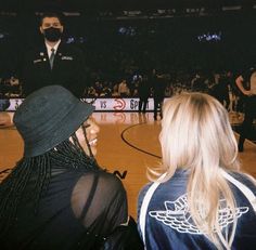 two women sitting on a basketball court with an air force officer standing behind them and people in the background