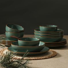 a set of teal green dishes with gold rims on a placemat next to pine needles