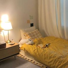 a bed with yellow sheets and pillows next to a night stand, lamp and window