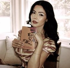 a woman sitting on a couch holding a book in front of her face and looking at the camera