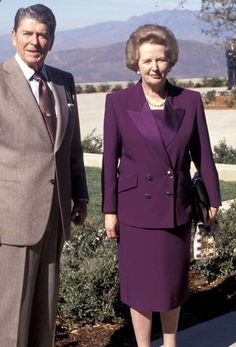 an older man and woman standing next to each other in front of some bushes with mountains in the background