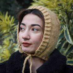 a woman wearing a knitted hat in front of some plants and bushes with her eyes closed