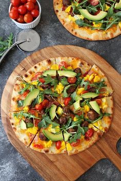 two pizzas sitting on top of a wooden cutting board next to a bowl of tomatoes and avocado