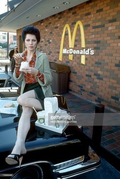 a woman sitting on the hood of a car eating mcdonald's food and drinking