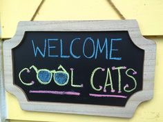 a welcome sign hanging from the side of a yellow building with chalk writing on it