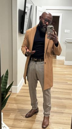 a man taking a selfie in the mirror with his cell phone while wearing a tan coat