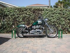 a green motorcycle parked in front of a hedge on a brick sidewalk next to a fence