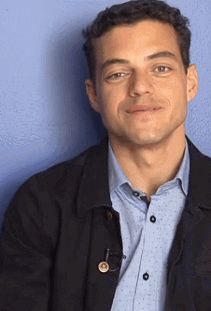 a man in a blue shirt and black jacket posing for a photo against a blue wall