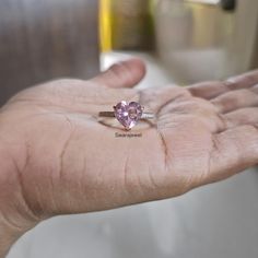 a person's hand holding a ring with a pink heart shaped stone on it