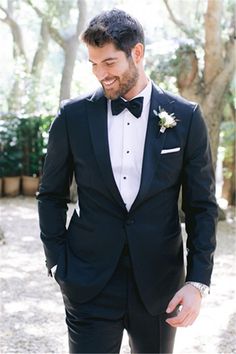 a man in a tuxedo smiles at the camera while standing under some trees