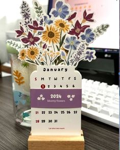 a desk calendar with flowers in it on a wooden stand next to a computer keyboard