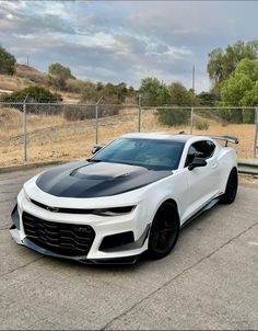 a white chevrolet camaro with black stripes parked in front of a chain link fence