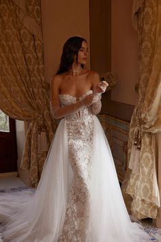 a woman in a white wedding dress standing next to a window with curtains and drapes