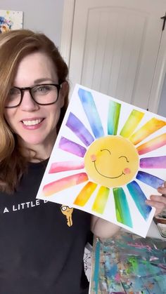 a woman holding up a painting with a smiley face on it and the sun painted in rainbow colors