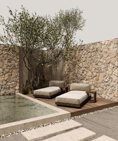 two lounge chairs sitting next to a pool in front of a stone wall and tree