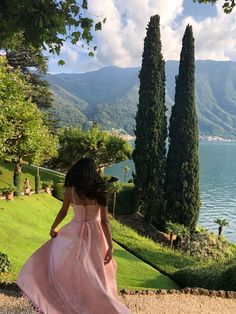 a woman in a pink dress is walking towards some trees and water with mountains in the background