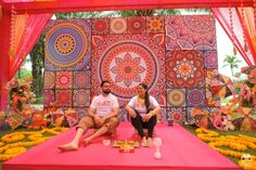 two people sitting on a pink mat in front of a wall with flowers and decorations