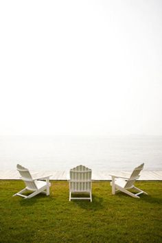 three white lawn chairs sitting on top of a lush green field next to the ocean