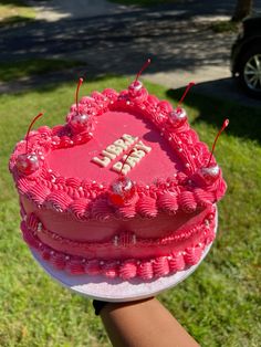 a pink heart shaped cake sitting on top of a grass covered field next to a person's hand