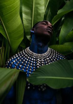 a man with his face covered in blue and silver jewelry surrounded by green leaves, looks up into the sky