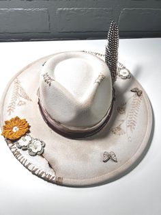 This wide-brim hat features a soft beige tone with intricate hand-stitched details along the crown and brim. Decorated with a mix of crochet flowers in warm earth tones and a single feather, it exudes a rustic, nature-inspired elegance. Vintage Beige Hat For Festival, Vintage Beige Festival Hat, Artisan Wide Brim Beige Hat, Artisan Beige Wide Brim Hat, Artisan Brimmed Beige Hat, Rustic Brimmed Hat For Spring, Rustic Short Brim Hat For Spring, Artisan Beige Brimmed Hat, Rustic Wide Brim Fedora For Spring