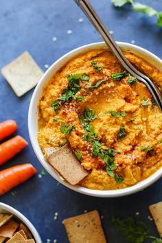 a white bowl filled with hummus and carrots next to crackers on a blue surface