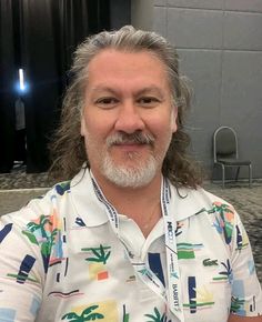 a man with long gray hair wearing a hawaiian shirt and smiling at the camera while sitting in front of a table