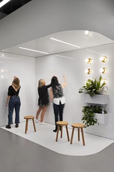 three women standing in front of a white wall with plants and stools on it