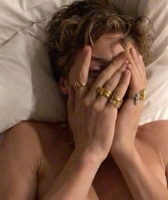 a shirtless woman covers her face while laying in bed with two gold rings on her hands